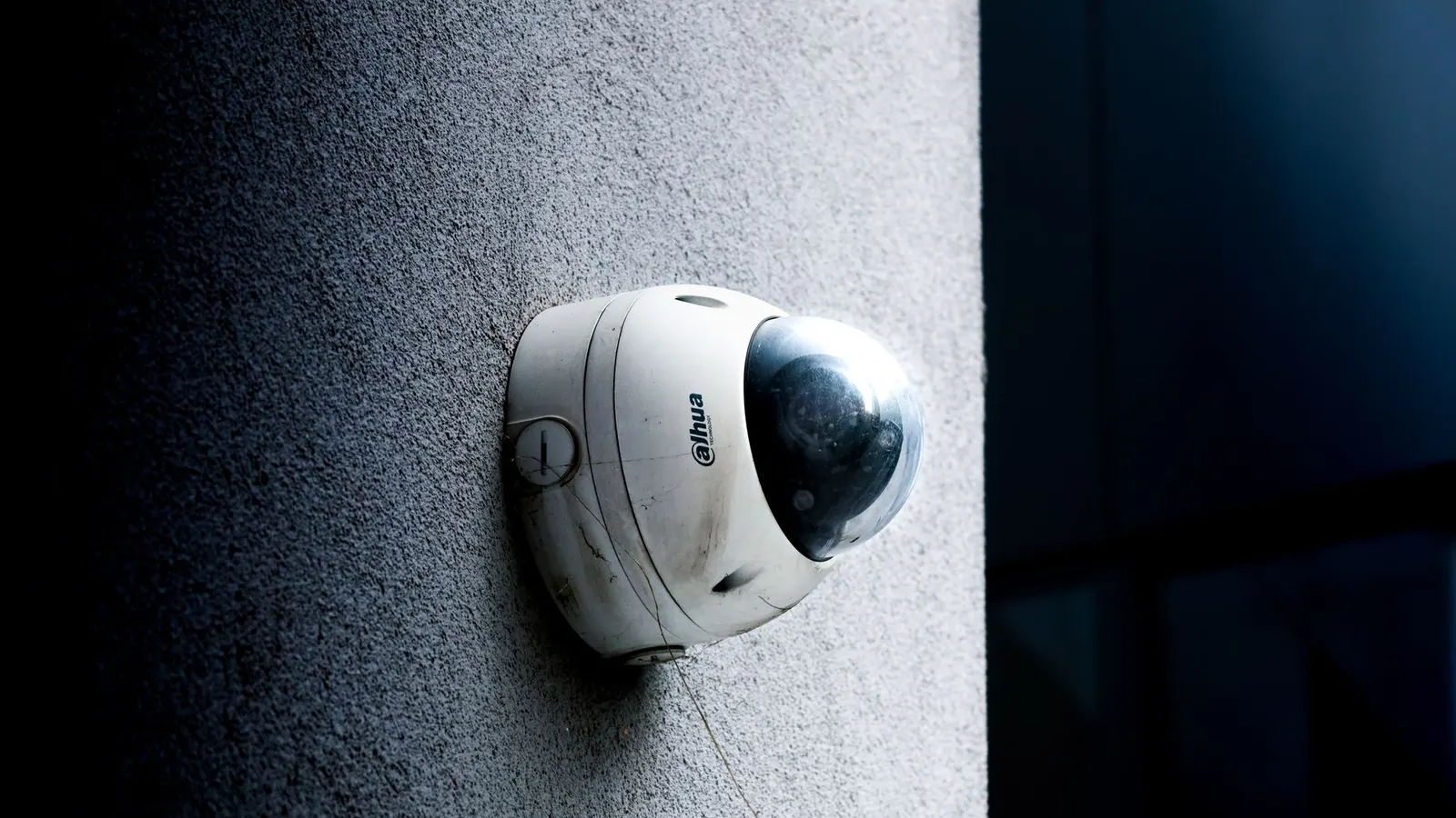 A white dome security camera bolted to a plain concrete wall, seen from below