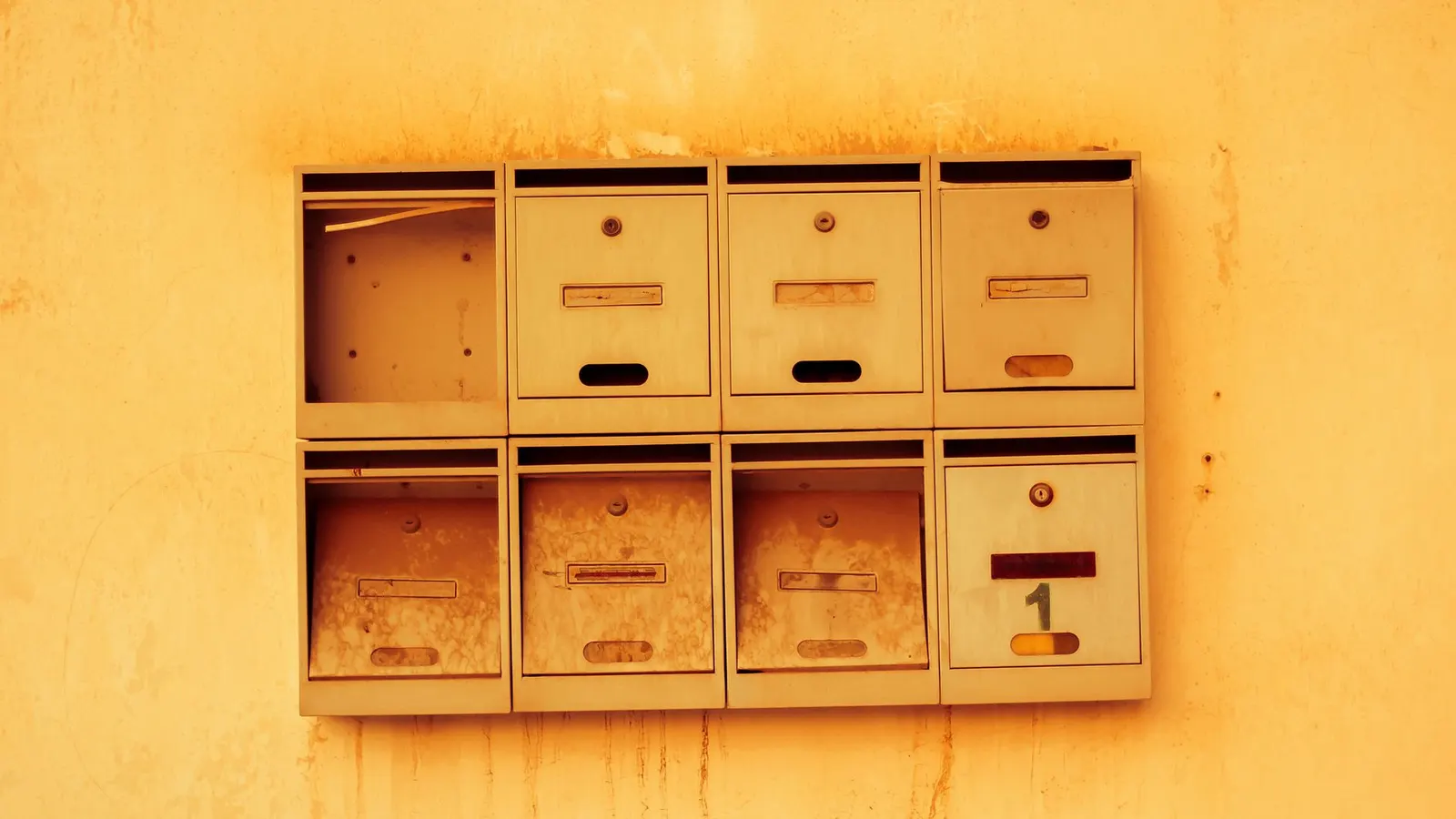 Eight weathered metal letterboxes in two rows on a yellow wall, their name slots blank and the door missing from one of them
