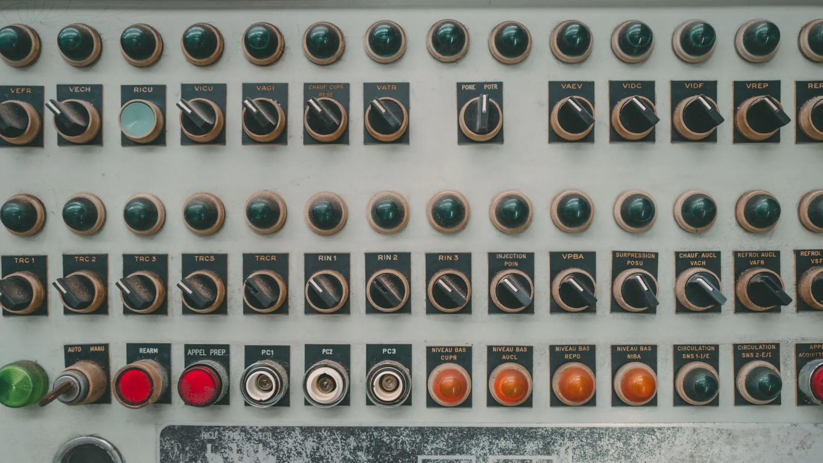Rows of selector switches and unlit indicator lamps on a grey industrial control panel, each labelled with a short abbreviation