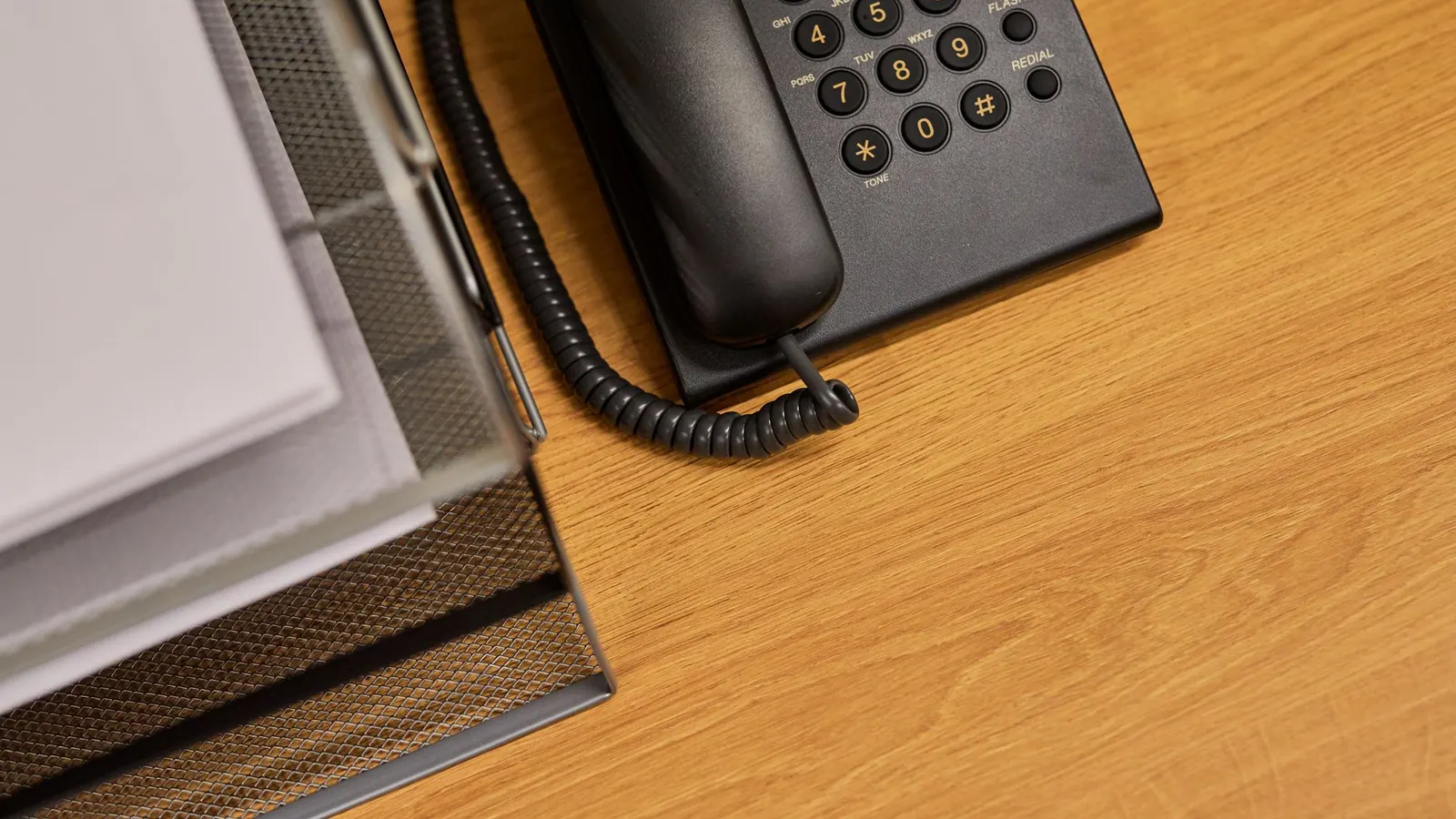 A black corded desk telephone beside a wire tray of documents on a wooden desk