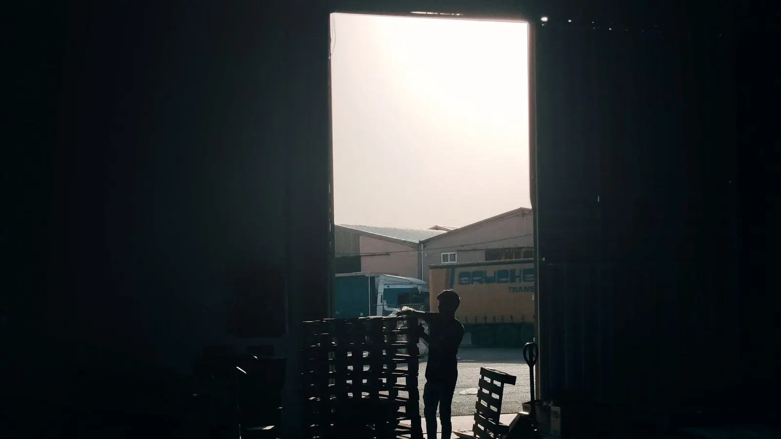 A worker silhouetted in a loading bay doorway, stacking crates against the bright daylight of the yard outside