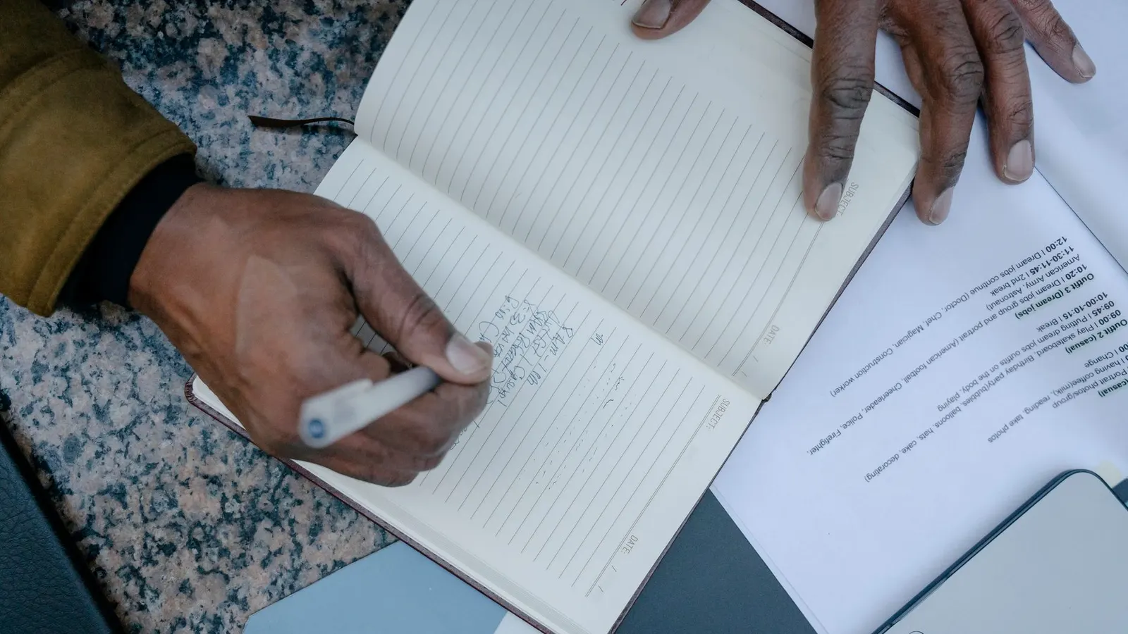 Looking down on a hand writing in a ruled notebook, with printed documents and a pen spread across the desk around it