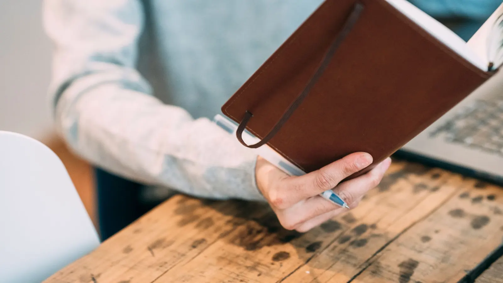 A man at a wooden office table turning the pages of a brown leather planner, pen in hand