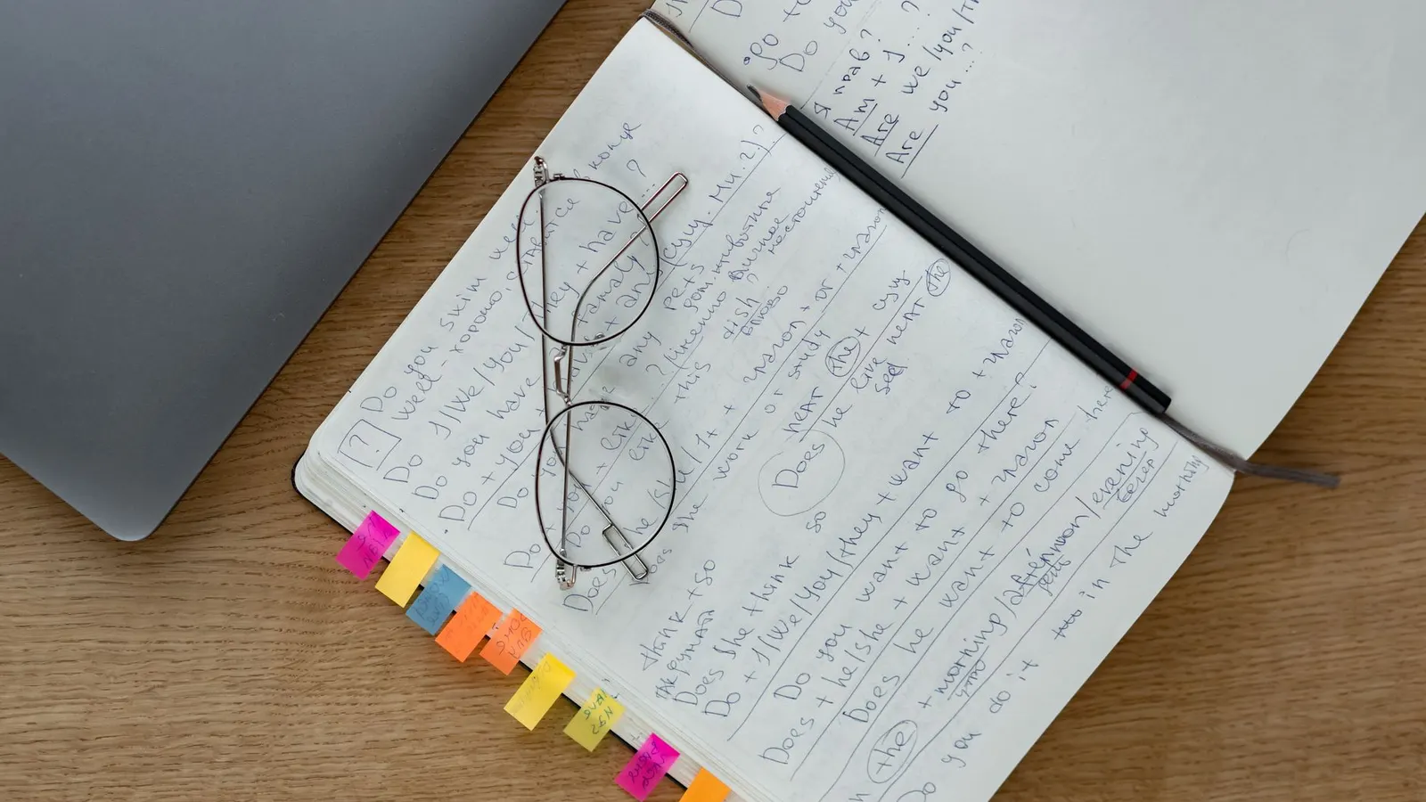 An open notebook covered in handwritten notes, with reading glasses and a pencil resting on it beside a laptop