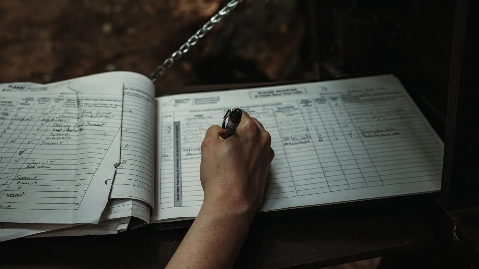 A hand signing a ruled register book chained to its stand, the columns beside it already filled with earlier entries in different handwriting