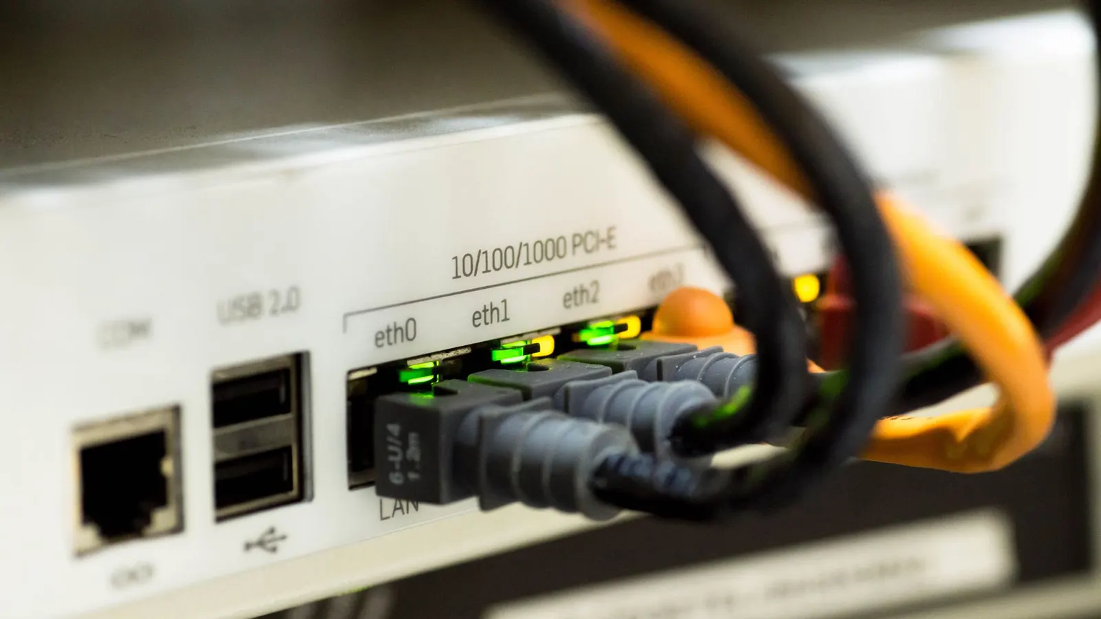 Close-up of the back of a white network device, with grey and orange cables plugged into labelled ethernet ports and the link lights showing green