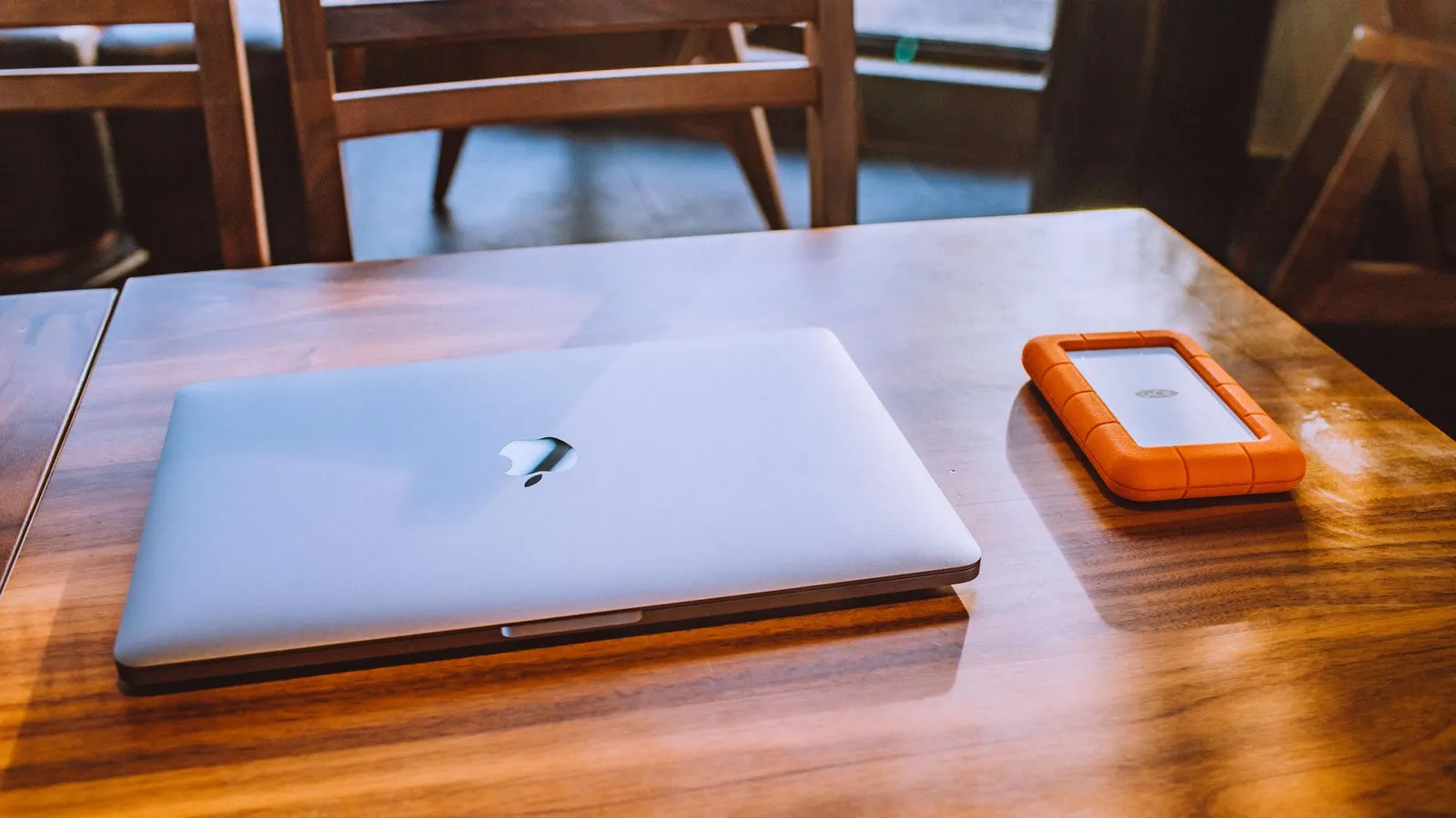 An open laptop on a wooden desk with a rugged portable hard drive plugged in beside it