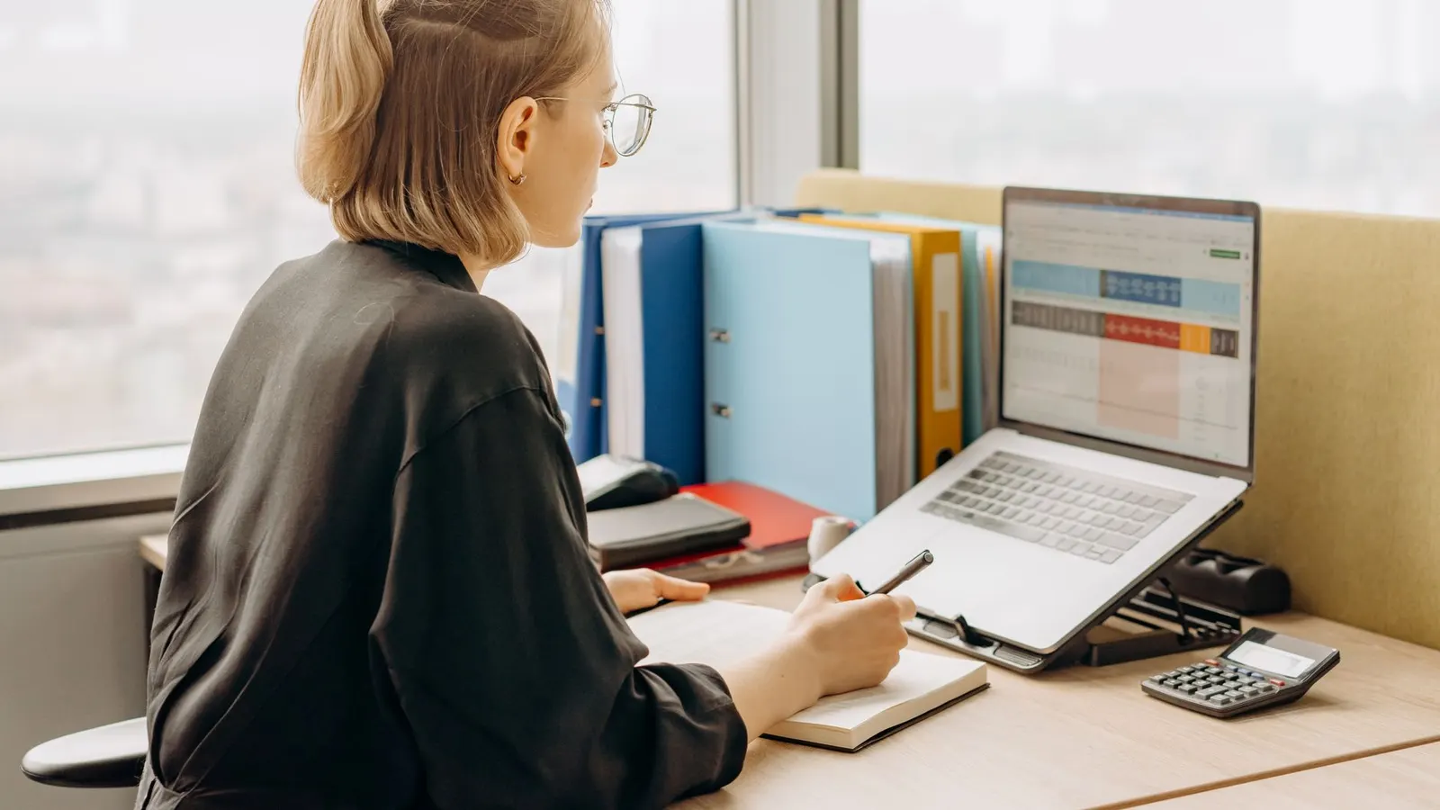 A woman at an office desk working through a spreadsheet on her laptop, seen from the side