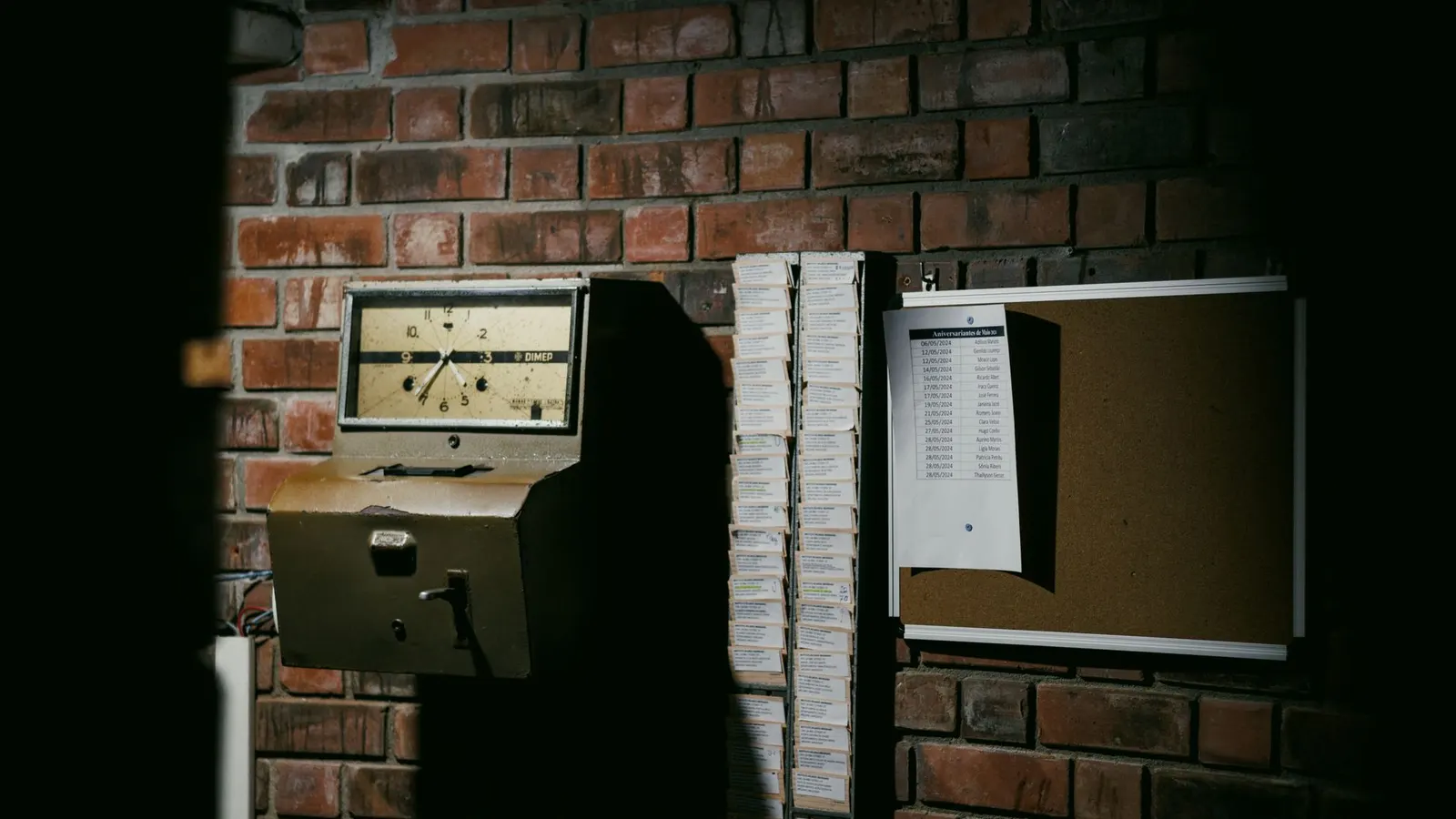 A wall-mounted clocking-in machine beside racks of timecards, with a printed list of names pinned to a noticeboard on a brick wall
